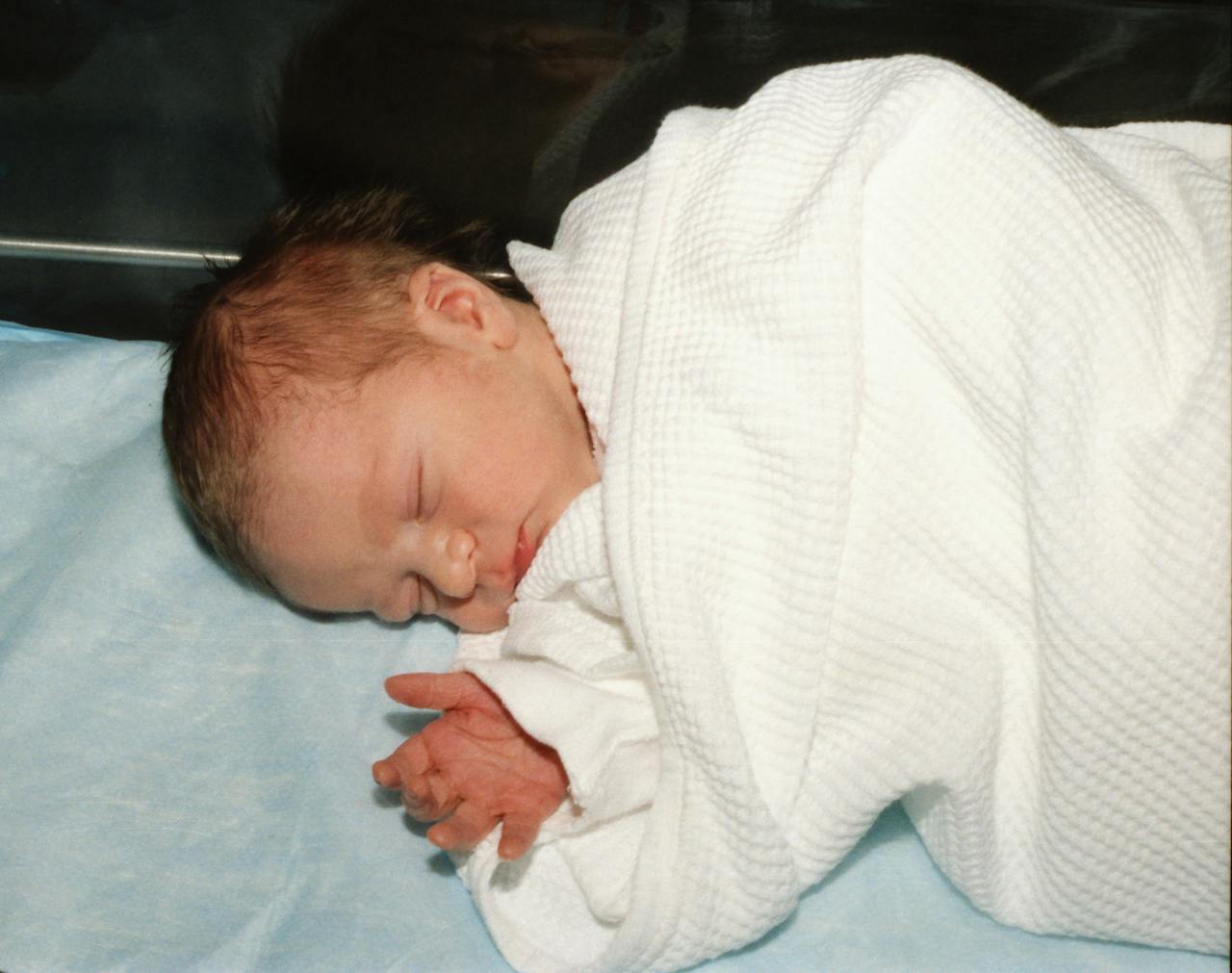 About A serene newborn baby peacefully sleeping, wrapped in a white hospital blanket on a light blue sheet.