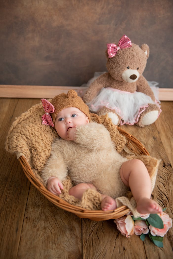 About Cute baby dressed in a bear outfit lying in a basket with a plush teddy bear nearby.