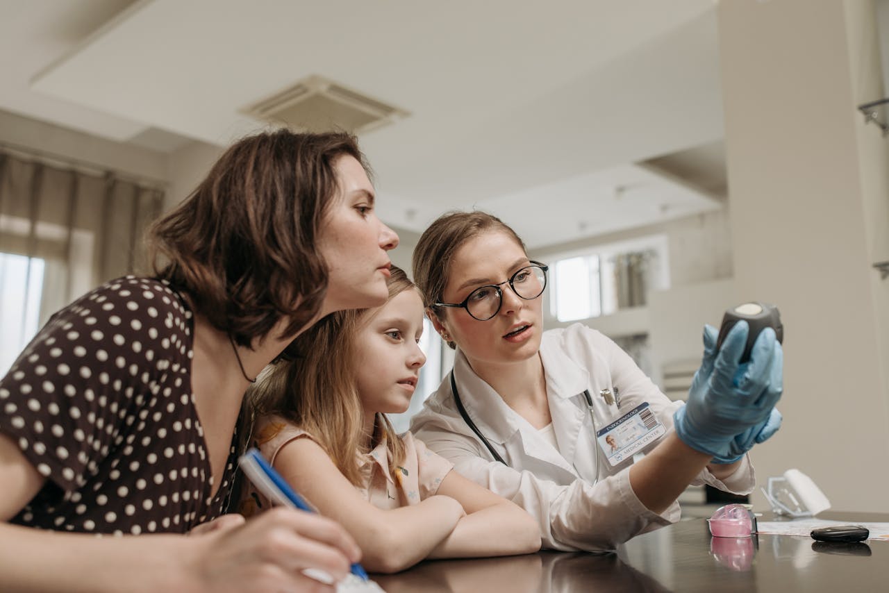 About A doctor explains a glucometer to a mother and child in a clinic setting indoors.